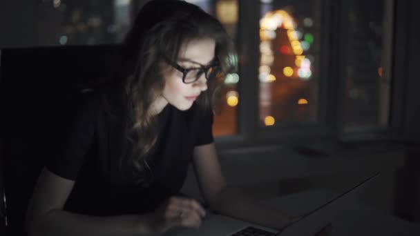 Πορτρέτο του ένα νεαρό ελκυστικό επιχειρηματικό γυναίκα που εργάζεται σε ένα φορητό υπολογιστή αργά το βράδυ στο γραφείο. κορίτσι με ένα επαγγελματικό κοστούμι και γυαλιά στο παρασκήνιο από τα φώτα της πόλης τη νύχτα — Αρχείο Βίντεο