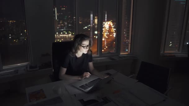 Gerente trabalha até tarde no escritório. jovem empresária trabalhando em um laptop tarde da noite contra as luzes da cidade noturna . — Vídeo de Stock