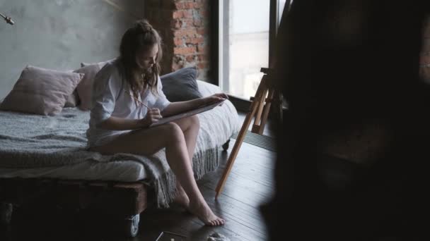 女の子のアーティストは、シャツのベッドに座って絵を描きます。画家は午前中にホームスタジオで働く. — ストック動画