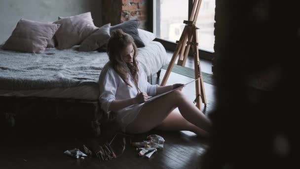 Painter works in a home studio in the morning. girl artist paints a picture of sitting on the bed in the shirt. — Stock Video