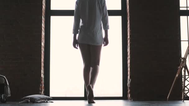 Porträtt av en smal flicka på bakgrunden av ett ljust fönster. flicka i skjorta går på tå — Stockvideo