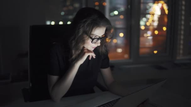 OSVČ, práci v kanceláři. dívka v brýlích funguje na notebooku na pozadí světla města — Stock video