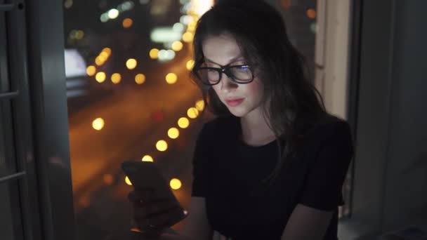 Retrato de una niña a la luz de la pantalla del teléfono inteligente. chica utiliza un teléfono móvil contra el fondo de la ciudad de la noche — Vídeo de stock