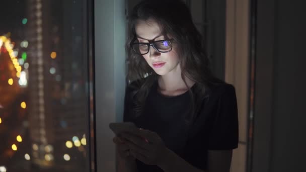 Reflejo de la pantalla con gafas. chica utiliza un teléfono inteligente en la oscuridad. retrato de una joven sobre el fondo de la ciudad nocturna — Vídeo de stock