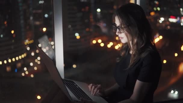 Praca w biurze, siedząc na parapecie przez duże okno kobieta młody na własny rachunek. biznes kobieta pracuje na laptopie późno w nocy w biurze na tle miasta świateł w nocy. — Wideo stockowe