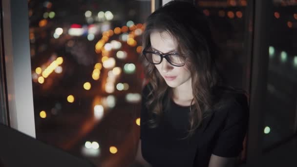Vértes portré egy vonzó lány, működő-ra egy laptop késő este. elmélkedés a monitor a szemüveg. fiatal nő dolgozik egy számítógépen, közepette az éjszakai város fényei. — Stock videók