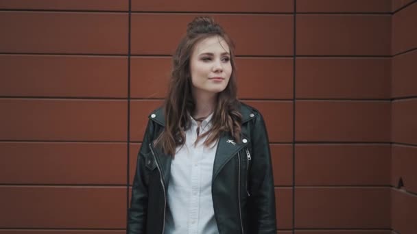 Hipster-Mädchen in Lederjacke vor dem Hintergrund moderner Architektur. Mädchen blickt und lächelt in die Kamera. — Stockvideo