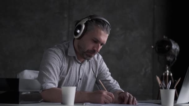 Arquitecto trabaja con documentos y dibujos en la oficina. hombre de mediana edad escucha música con auriculares en la oficina . — Vídeos de Stock
