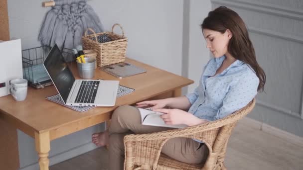 Молода жінка залишає журнал, сидячи за столом вдома — стокове відео