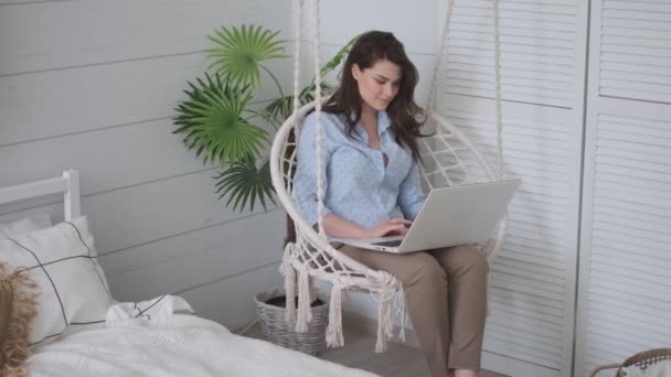 Kız rahat bir asılı sandalyede kucağında bir dizüstü bilgisayar ile oturuyor. evde çalışan genç kadın serbest çalışan. — Stok video