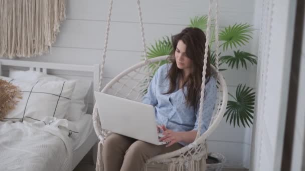 自由职业者在家里工作，坐在舒适的吊椅上。女孩工作在笔记本电脑躺在她的腿上. — 图库视频影像