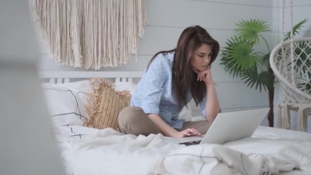 女の子はベッドに座っている間ラップトップを使用します。自宅でカジュアルで魅力的な若い女性の肖像画. — ストック動画
