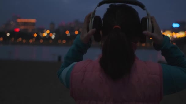 Mädchen setzt Kopfhörer auf und joggt durch die nächtliche Stadt. — Stockvideo