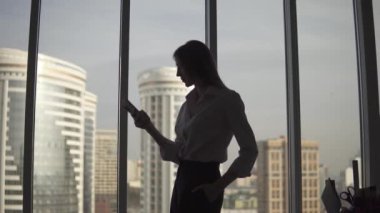 Büyük pencere ve gökdelen çerçevede genç bir kadın silueti. Kız bir akıllı telefon kullanır.