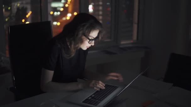 Retrato de una joven atractiva mujer de negocios que trabaja en una computadora portátil tarde en la noche en la oficina. chica en un traje de negocios y gafas en el fondo de las luces de la ciudad de la noche — Vídeo de stock