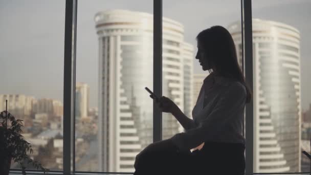 Силуэт девушки с телефоном в руках на фоне современных зданий. бизнес-леди пользуется смартфоном в офисе . — стоковое видео