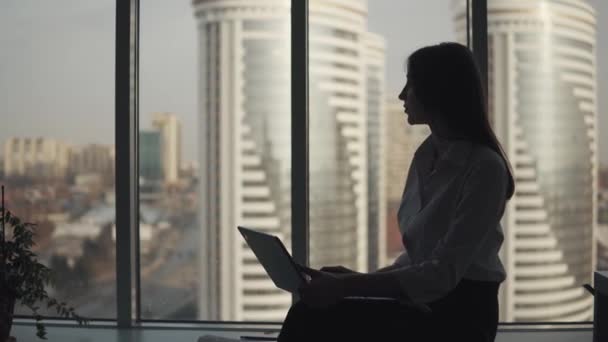 Retrato de una mujer de negocios que trabaja en un portátil en la oficina cerca de la ventana. silueta de una chica que trabaja hasta tarde sola . — Vídeos de Stock