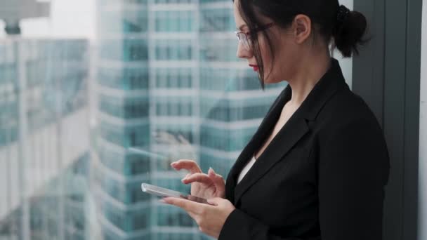 Mujer de negocios usando un teléfono celular mientras está de pie en un piso alto de un rascacielos cerca de una ventana . — Vídeos de Stock