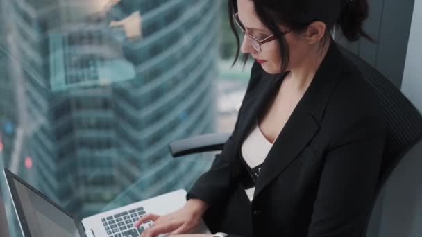 Portret młodej bizneswoman pracując na laptopie siedząc przez okno na wysokim piętrze wieżowca. — Wideo stockowe