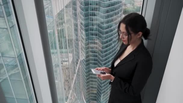 Mujer joven en ropa de negocios utiliza un teléfono inteligente mientras está de pie en un piso alto de un rascacielos — Vídeos de Stock