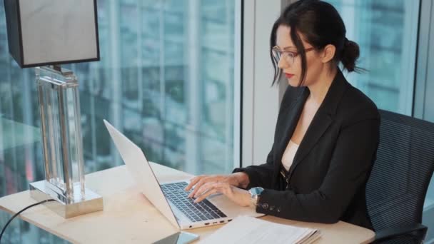Νεαρή γυναίκα που εργάζεται στο laptop κάθεται δίπλα στο παράθυρο στο γραφείο — Αρχείο Βίντεο