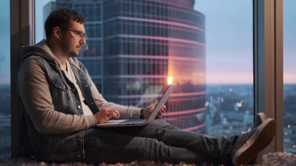 Portrait of a young man sitting thoughtfully behind a laptop by a window on a high floor — Stock Video