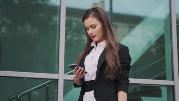 Портрет деловой женщины с мобильным телефоном в руках. Молодая женщина в деловом костюме пользуется смартфоном, стоя на улице — стоковое видео