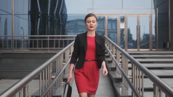 Magabiztos üzleti nő egy piros ruhában sétálni mellett egy üzleti központ. fiatal nő egy piros ruhában egy táskát a kezében. — Stock videók