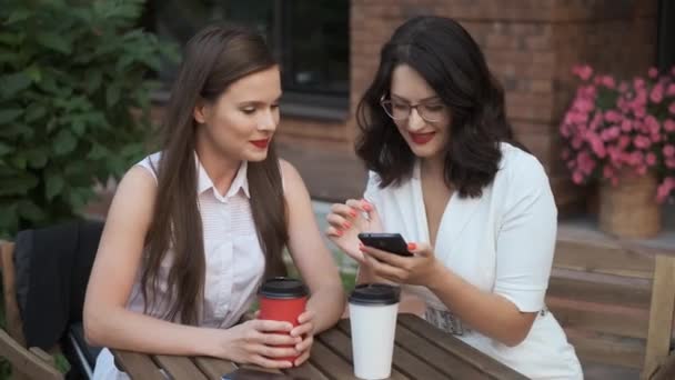 Zakelijke vrouwen zitten in een zomercafé en praten. meisje toont iets aan haar vriend op het scherm van de smartphone. — Stockvideo