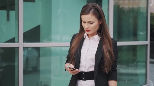 Молодая деловая женщина просматривает информацию на своем смартфоне и уверенно идет по улице — стоковое видео