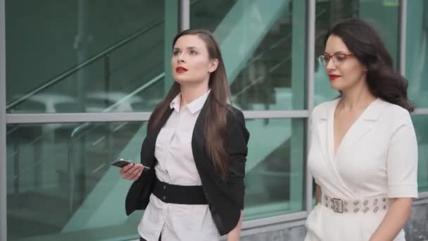 Las mujeres de negocios caminan junto a un moderno edificio de negocios. dos mujeres atractivas están hablando sobre la marcha . — Vídeo de stock