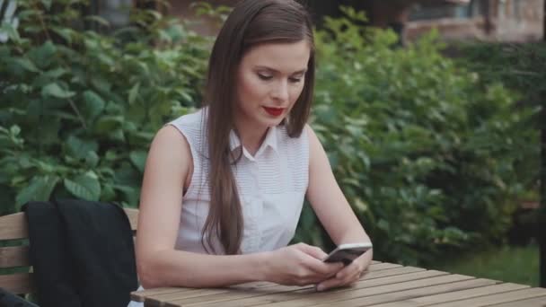 Junge Geschäftsfrau sitzt in einem Sommercafé und chattet mit einem Smartphone — Stockvideo