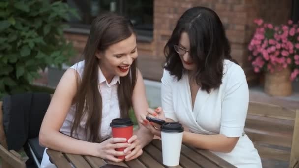 Молода жінка показує своєму другові щось на телефоні. дві бізнес-леді сидять за столом в літньому кафе і весело спілкуються — стокове відео