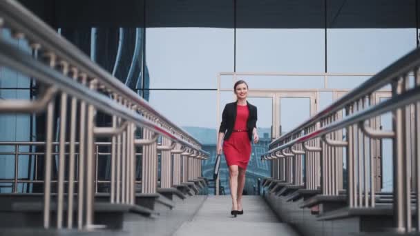 Zelfverzekerde zakenvrouw in een rode jurk loopt tegen de achtergrond van moderne gebouwen. Portret van een jonge vrouw in een jasje en het dragen van een aktetas in haar handen — Stockvideo