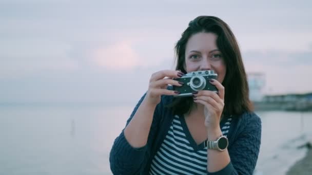 Νεαρή γυναίκα παίρνει φωτογραφίες σε μια vintage κάμερα δίπλα στον ωκεανό. πορτρέτο ενός κοριτσιού με μια ρετρό κάμερα — Αρχείο Βίντεο