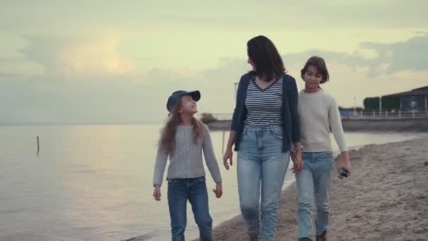 Madre joven camina con sus hijos a la orilla del mar al atardecer. la familia pasa tiempo al aire libre . — Vídeos de Stock