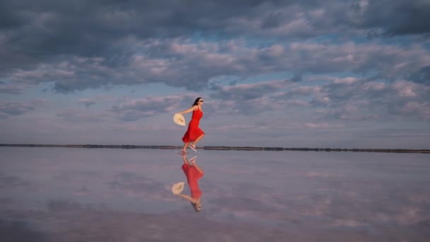 Κορίτσι με ένα φόρεμα που φτερουγίζει διασχίζει μια αλμυρή λίμνη. τα σύννεφα αντανακλώνται στο νερό — Αρχείο Βίντεο