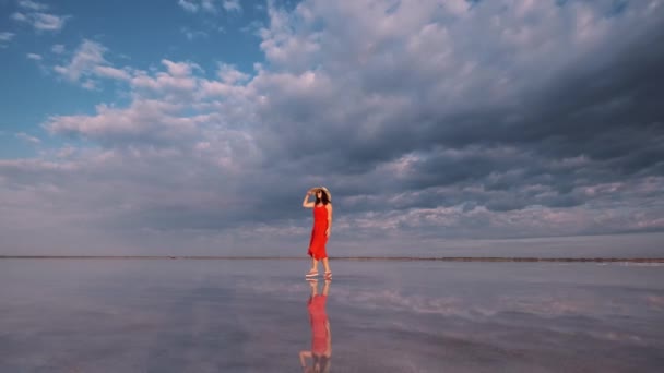 Turista mulher em um vestido acenando caminha ao longo de um lago de sal no qual o céu se reflete — Vídeo de Stock