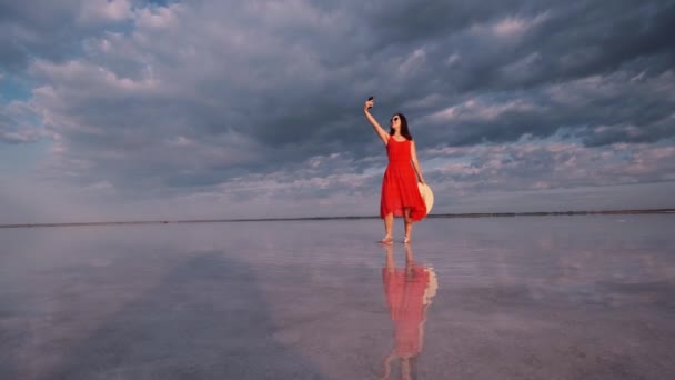 Jeune femme prend un selfie tout en se tenant sur le rivage d'un lac rose dans lequel le ciel se reflète — Video