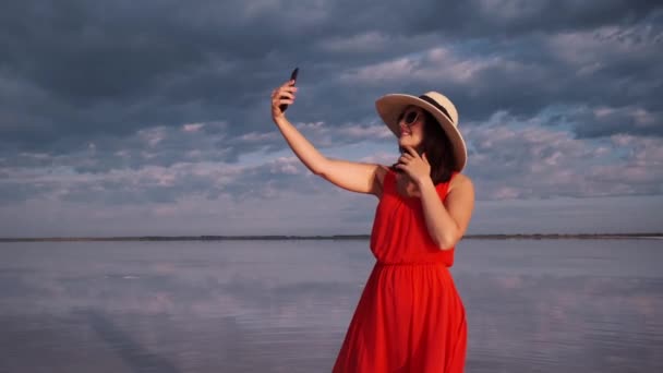 Junge Frau macht ein Selfie an einem wunderschönen, ungewöhnlichen Ort. Mädchen in rotem Kleid, Hut und Sonnenbrille auf einem rosa See. — Stockvideo