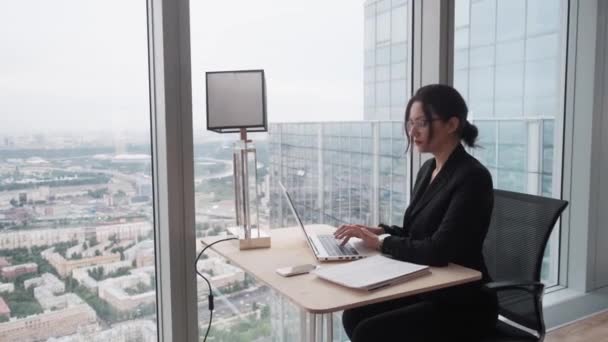 オフィスの窓のそばに座ってコンピュータで働くビジネスウーマン。職場でのビジネス服装の若い女性の肖像. — ストック動画