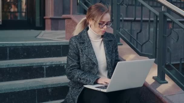 Jovem mulher em um casaco trabalha em um laptop enquanto sentado nos passos — Vídeo de Stock