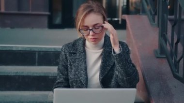 Gözlüklü ve ceketli çekici kız dışarıda dizüstü bilgisayar kullanıyor.