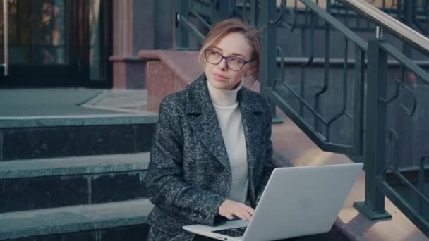 Mujer joven en gafas utiliza un ordenador portátil al aire libre. chica en un abrigo se sienta en las escaleras — Vídeos de Stock