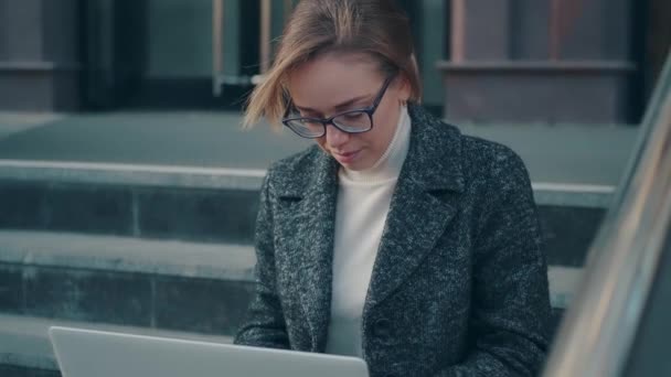 Jovem mulher trabalhando em um laptop ao ar livre. menina em um casaco senta-se nos passos — Vídeo de Stock