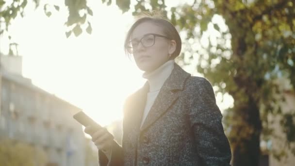 Γυναίκα χρησιμοποιεί ένα smartphone σε εξωτερικούς χώρους. πορτρέτο ενός κοριτσιού με γυαλιά και παλτό — Αρχείο Βίντεο