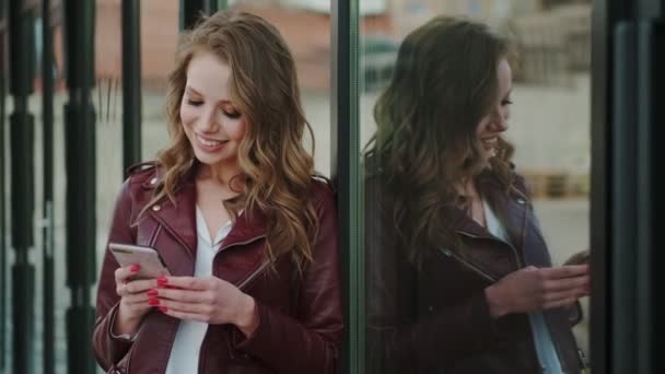 Chica atractiva con una sonrisa encantadora utiliza un teléfono inteligente y sonríe alegremente — Vídeos de Stock