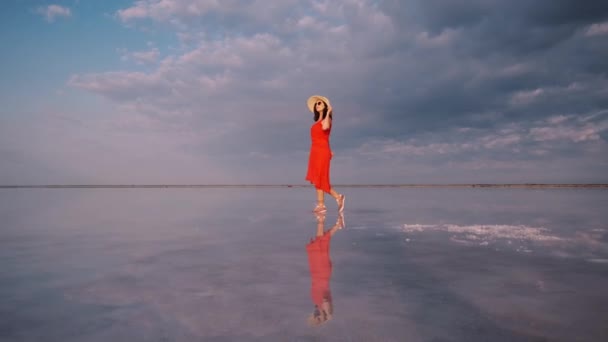 Menina em um vestido fluttering caminha ao longo de um lago de sal rosa em que o céu é refletido — Vídeo de Stock