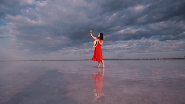 Νεαρή γυναίκα παίρνει μια selfie, ενώ στέκεται στην ακτή μιας ροζ λίμνης στην οποία ο ουρανός αντανακλάται — Αρχείο Βίντεο