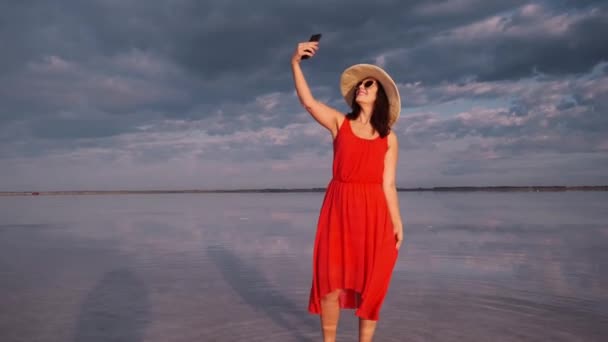 Jonge vrouw neemt een selfie in een prachtige ongebruikelijke plaats. meisje in een rode jurk, muts en zonnebril op een roze meer. — Stockvideo